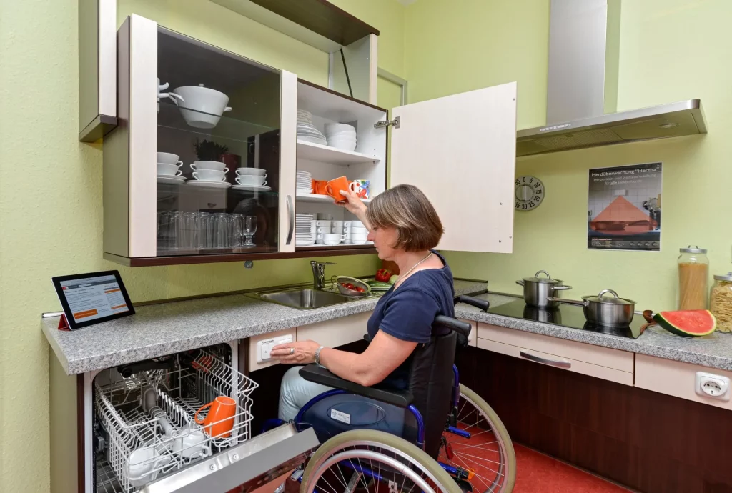 Frau im Rollstuhl, die sich die Hängeschränke per Knopfdruck herunterfahren lässt, um Geschirr aus der Spülmaschine in den Schrank einzuräumen