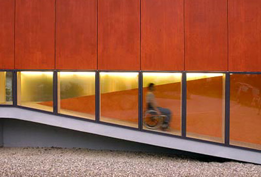 Person im Rollstuhl, die hinter einem Milchglasfenster im Treppenhaus eines Gebäudes eine Rollstuhlrampe herunterfährt.