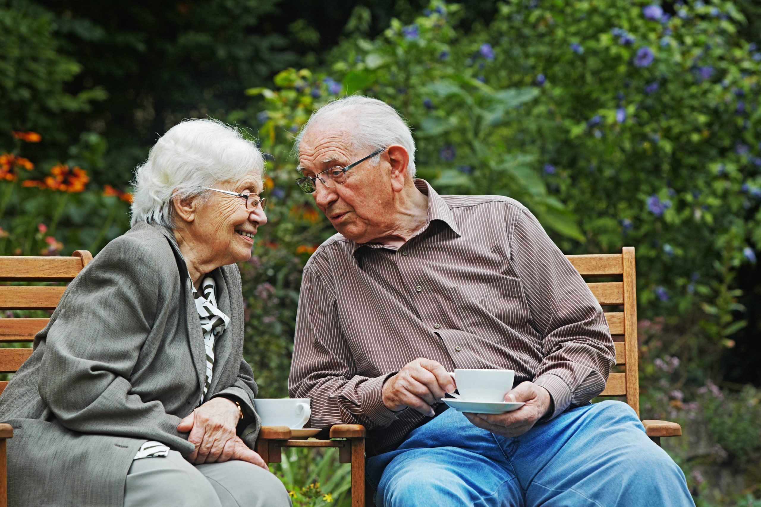 Ein älteres Ehepaar trinkt Kaffee auf einer Gartenbank und unterhält sich dabei.