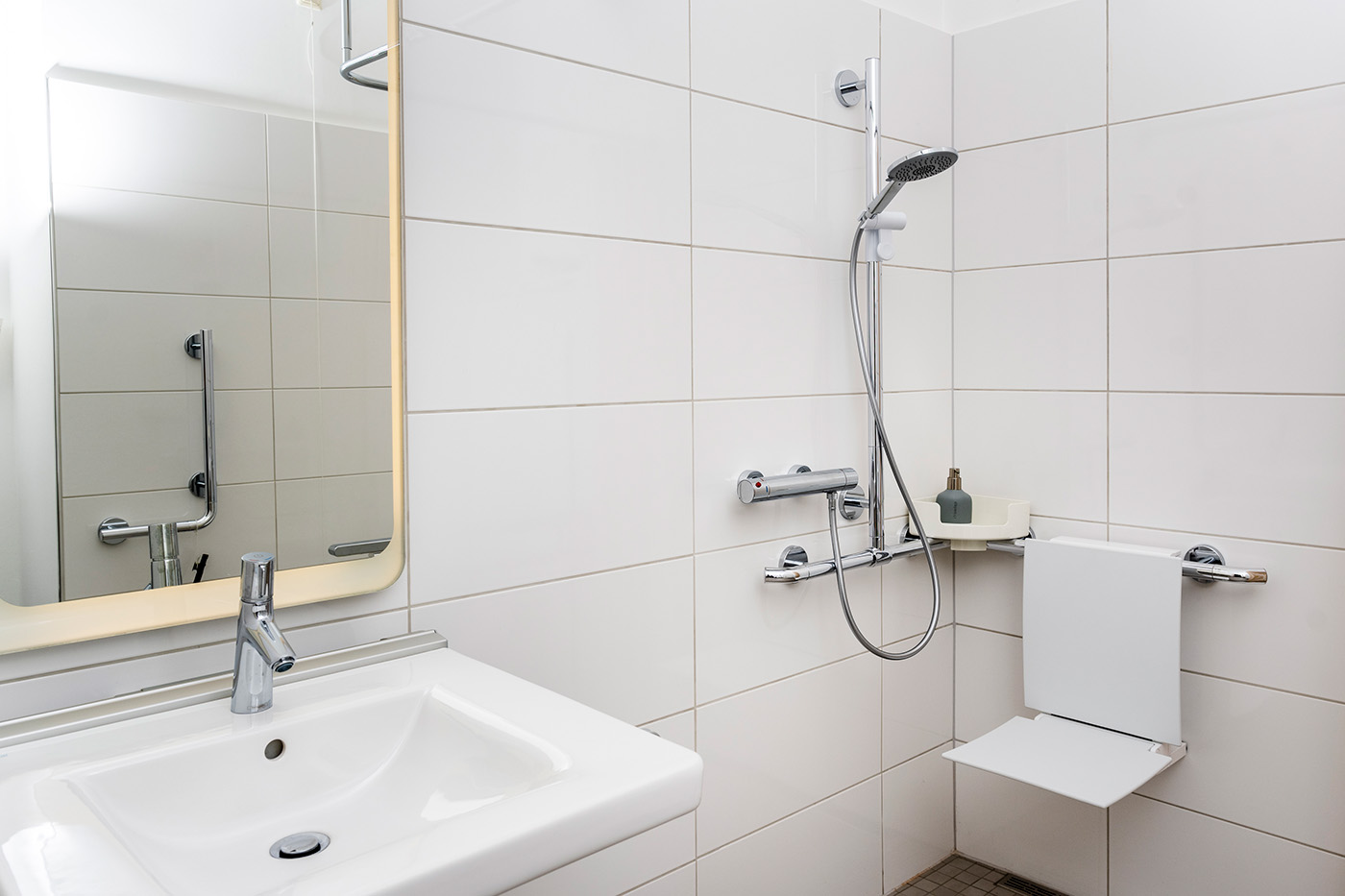 Barrierefreies Bad mit bodengleicher Dusche, deren Halterung als stabiler Haltegriff genutzt werden kann, daran montiertem Duschklappsitz und mit unterfahrbarem Waschtisch