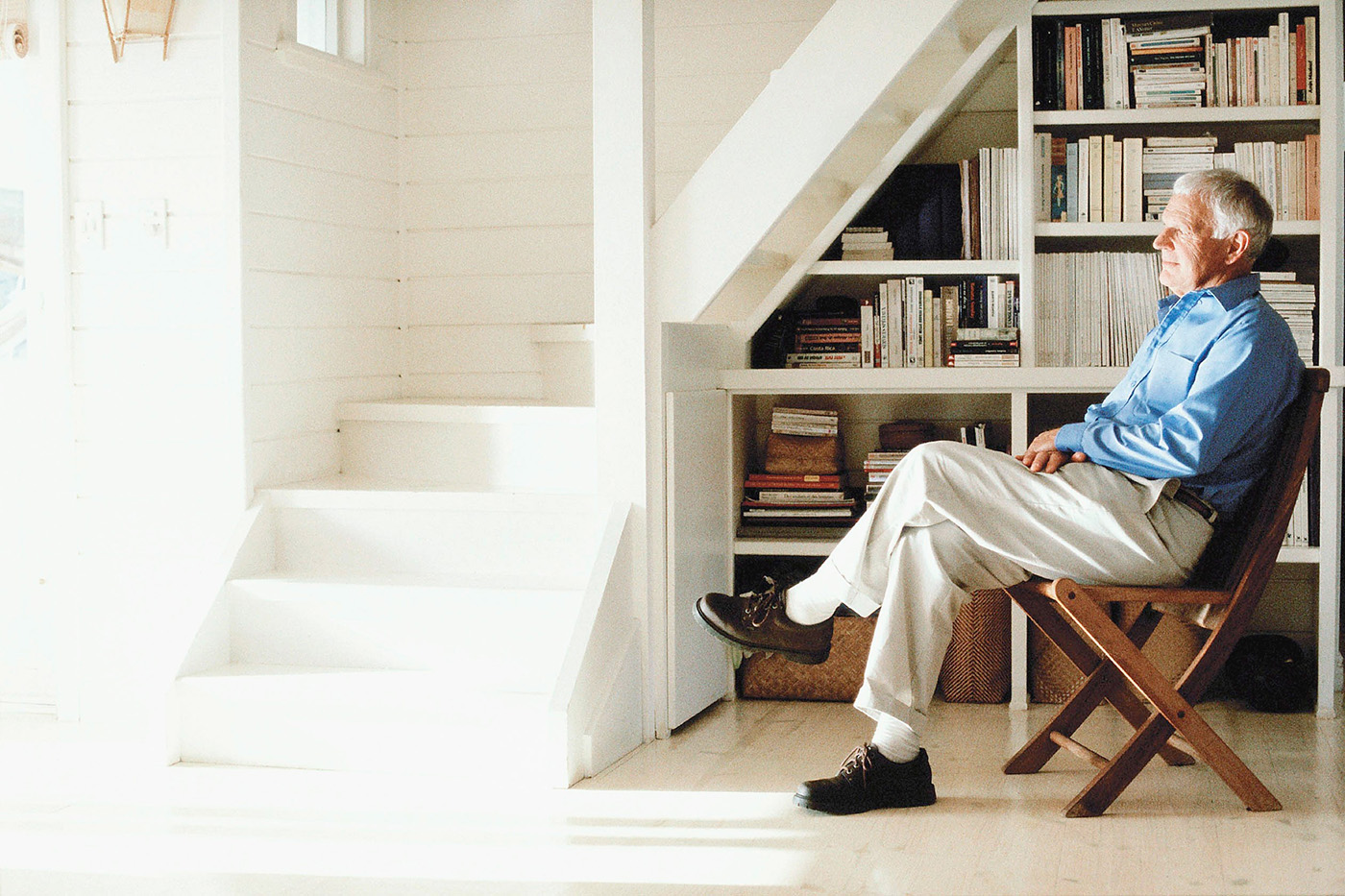 Älterer Herr sitzt in einem Sessel im Wohnzimmer neben einer nach oben führenden Treppe und schaut nachdenklich aus dem Fenster