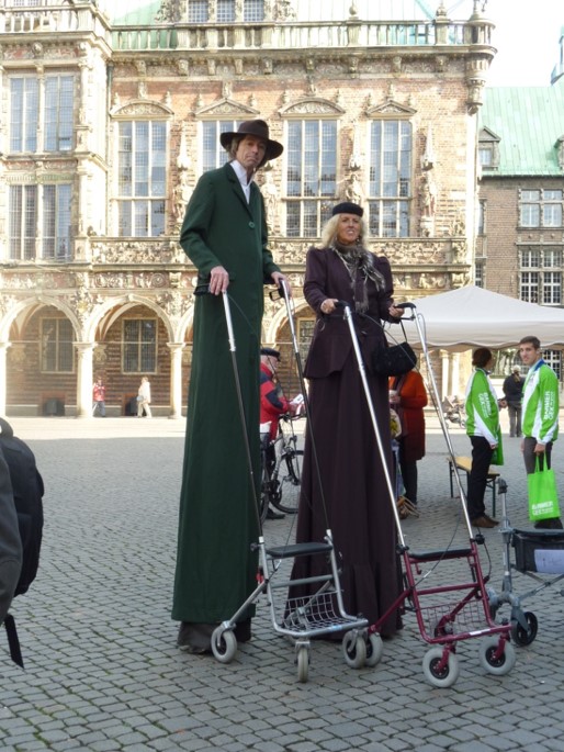 Stelzenläufer als Herr und Dame mit Hut gekleidet, mit  Rollatoren, die verlängerte Griffe haben, vor dem Bremer Rathaus