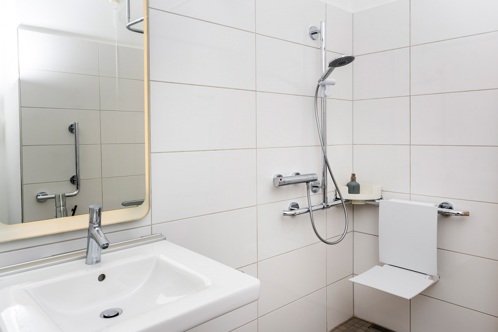 Barrierefreies Badezimmer mit einer bodengleichen Dusche mit Duschsitz, Haltemöglichkeiten und unterfahrbarem Waschbecken.