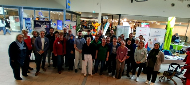Ein Gruppenbild des Netzwerkes Älter werden im Bremer Westen. Es sind mehr als 25 Personen zu sehen, die alle in die Kamera schauen. Im Hintergrund stehen verschiedene Messestände.