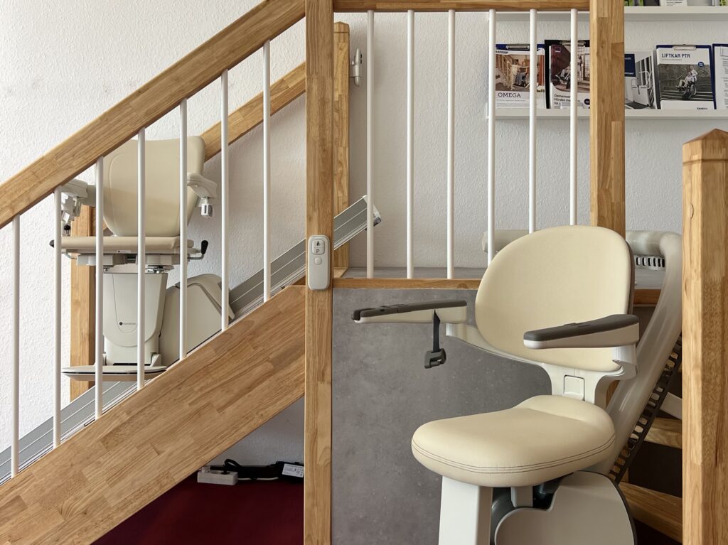 Zwei Treppenlifte an einem Holzpodest. Einer an einer kurvigen Treppe und einer an einer geraden Treppe.